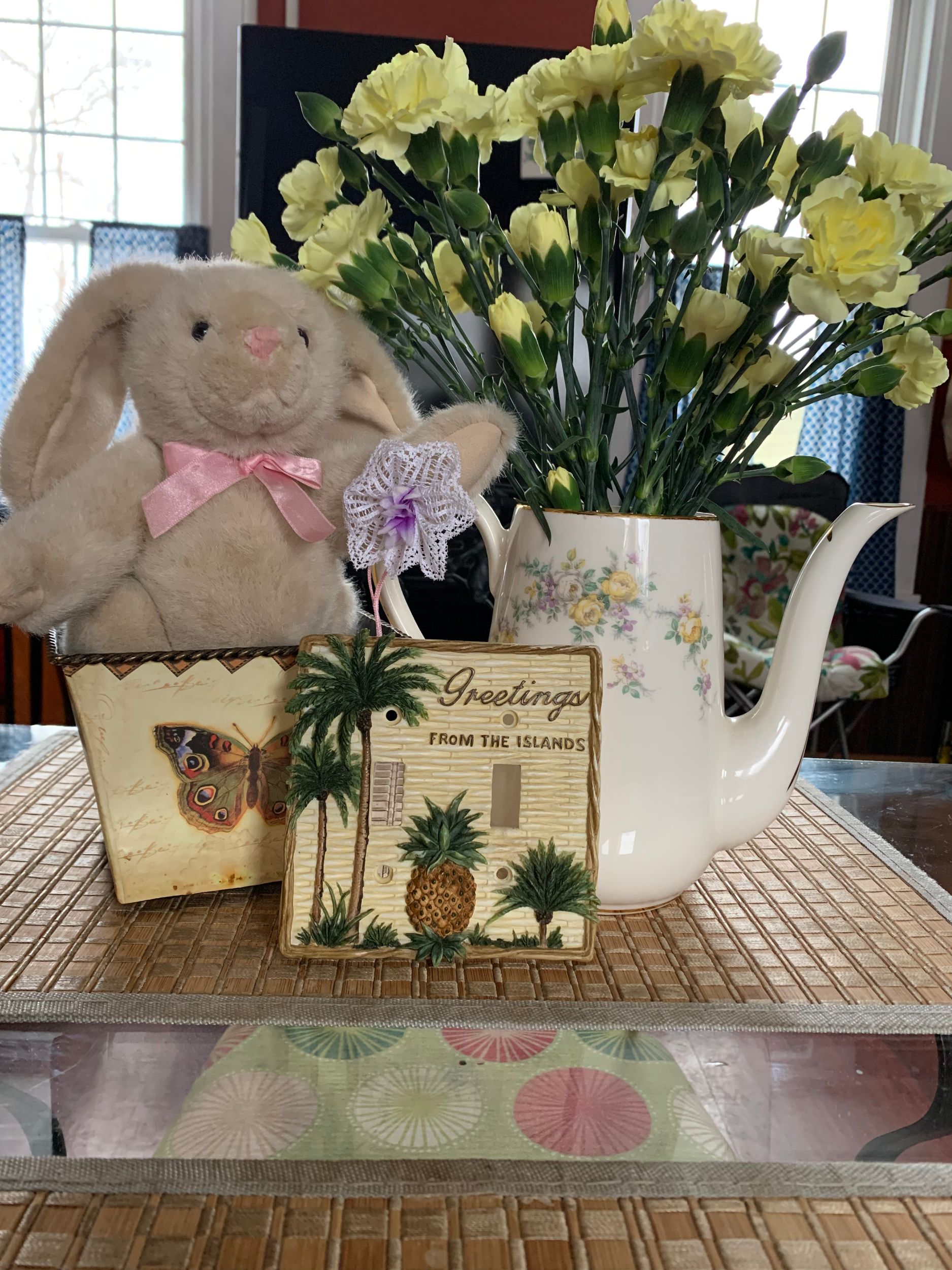Objects arranged on straw mat including plush rabbit in butterfly container, palm tree light switch, china coffee pot holding bunch of yellow carnations, with background windows showing snow falling